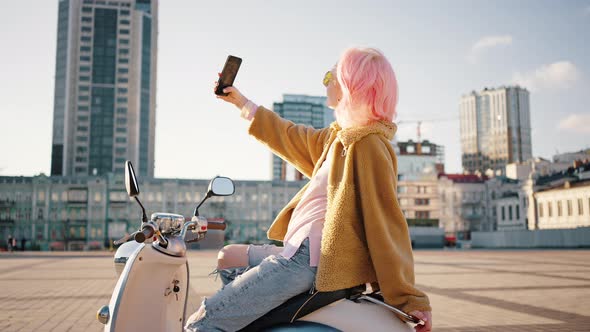 Young Stylish Woman with Pink Hair Making Selfie on Smartphone Outdoor Sitting on Scooter Tracking