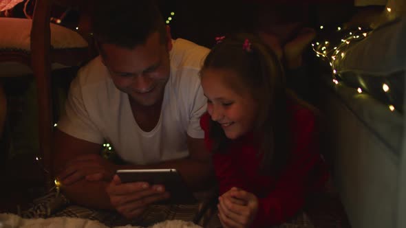 Caucasian father and daughter lying and using digital tablet under blanket fort during christmas