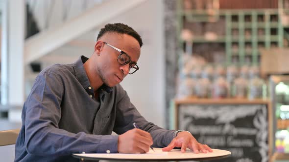 Serious Young African Man Writing on Paper in Cafe