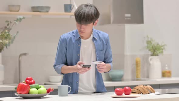 Asian Man Making Online Payment on Smartphone in Kitchen