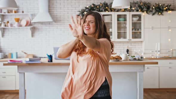 Cheerful Overweight Lady Listening to Music in Wireless Headphones and Actively Dancing at Kitchen