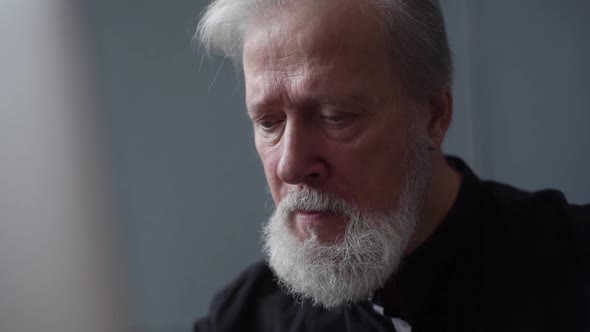 Closeup Face of Serious Bearded Senior Aged Businessman Working on Laptop at Workplace in Dark Home