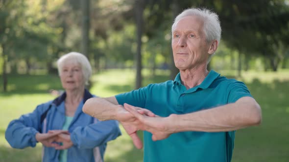 Confident Greyhaired Senior Man Stretching Hands Warming Up As Blurred Smiling Woman Exercising at