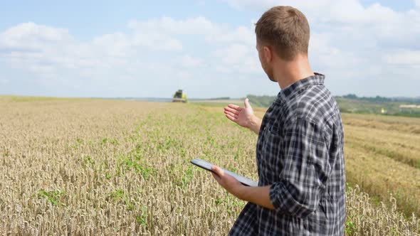 Farmer Using a Laptop and Holding Notebook with a Combine Harvester in a Wheat Field on Background