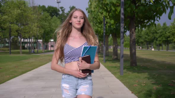 Young Attractive Woman Student Walks Outdoors with a Textbook and Notebook