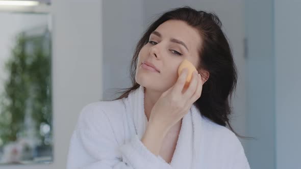 Attractive young brunette Woman in bathrobe  applying foundation with beauty blender at the bathroom