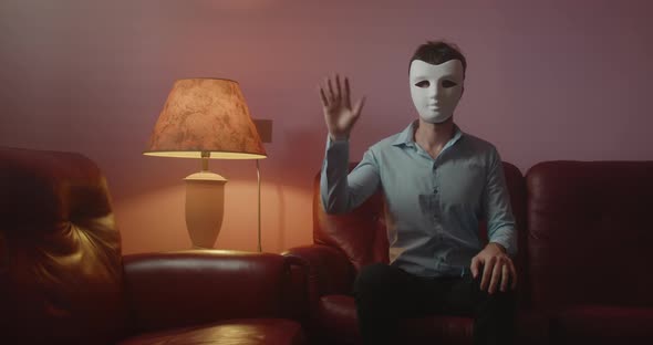 Anonymous man with mask greets with his hand while sitting on the red sofa at home