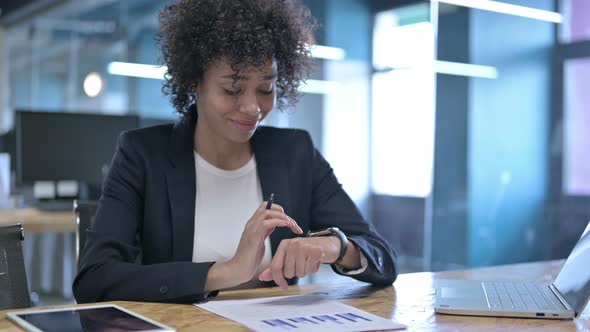 Cheerful African Businesswoman Using Smart Watch and Reading Documents in Office