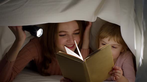Young Mother Reading Goodnight Story Fairytale to Child Daughter Under Duvet Blanket in Night Room