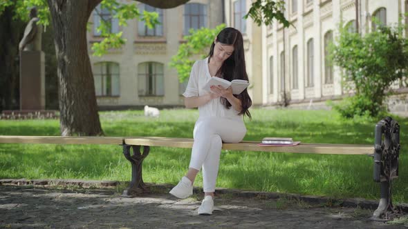 Wide Shot of Concentrated Brunette Woman in White Clothes Sitting on Bench on Sunny Day and Reading