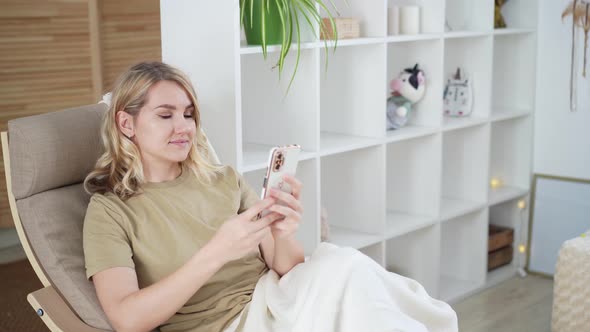 Cute young blonde woman uses phone while sitting at home in armchair Using technology concept