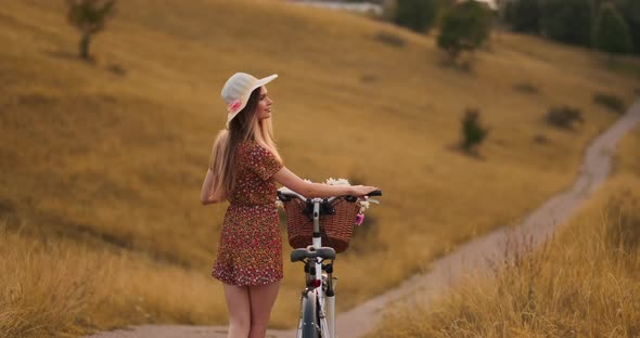 Smiling Happy Girl in Dress and Hat Riding Retro Bicycle in the Park and Looking at Camera