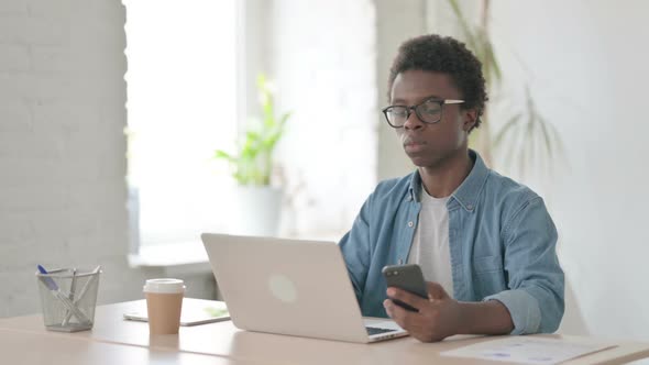 Young African Man Browsing Internet on Smartphone While Using Laptop in Office
