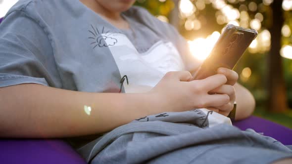 A Girl Plays or Texts on Social Networks with Friends Sitting on a Big Pillow in a City Park