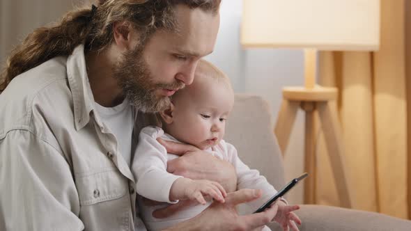 Caucasian Father Bearded Dad Holding Newborn Daughter Son Infant at Home Using Mobile App Browsing