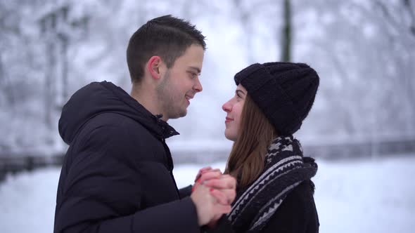 Portrait Cute Young Woman and Man Kissing in Winter Park Under Falling Snow. Happy Couple in Love