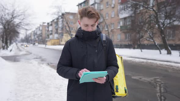 Male Student Messaging Online Closing Tablet and Walking Away with Yellow Food Delivery Bag