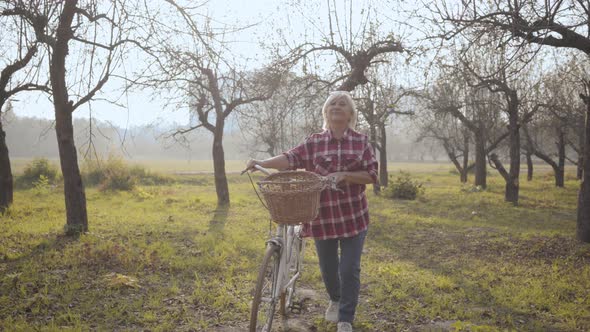 Smiling Mature Caucasian Woman in Casual Clothes Walking in the Foggy Park with Bicycle