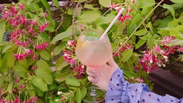 Woman Hand Holding Glass with Tropical Cocktail with Pink Flowers on Background