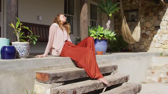 Happy caucasian woman sitting on steps, relaxing with eyes closed on sunny cottage terrace