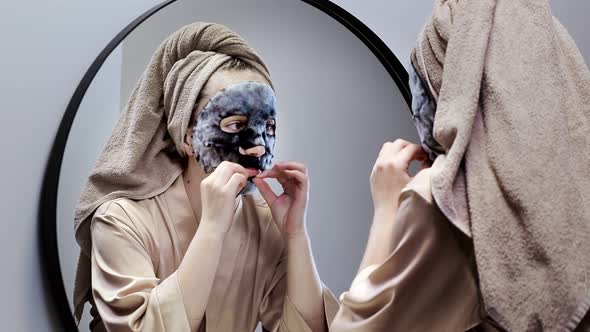 Young happy woman in towel making face mask and looking in mirror in stylish bathroom.