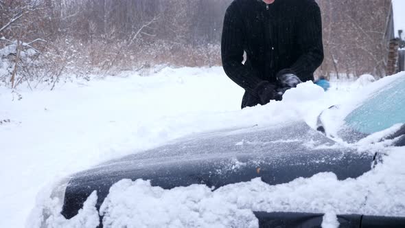 Man in a Black Jacket Cleans a Car From Snow on a Background of Trees