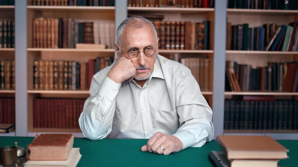 Elderly 70s Man Scientist Doctor Professor in White Shirt Sitting at Desk Workplace in Library