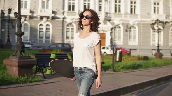 Smiling Attractive Woman in Sunglasses Walking in the Old City Street Holding Her Longboard in the