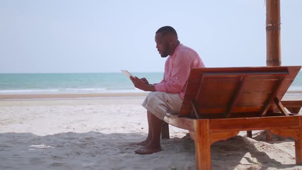 Mature African American man using a tablet while sitting on the beach.