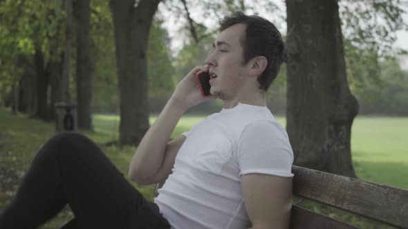 Side View of Young Millennial Brunette Man Sitting on Bench on Summer Park Alley Talking on the