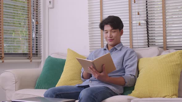 At home, a handsome young Asian businessman in casual clothing reads a book on the couch.