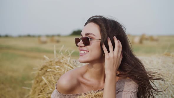 Happy Woman Rejoices at Haystack and Corrects Hair During Strong Wind in Field