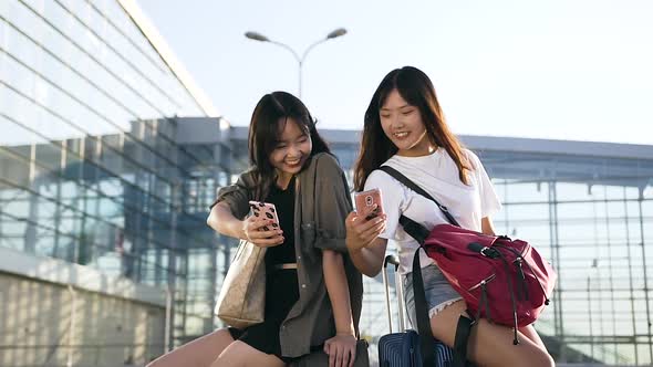 Two Charming Exuberant Asian Women which Sitting on their Suitcases and Using Smartphones