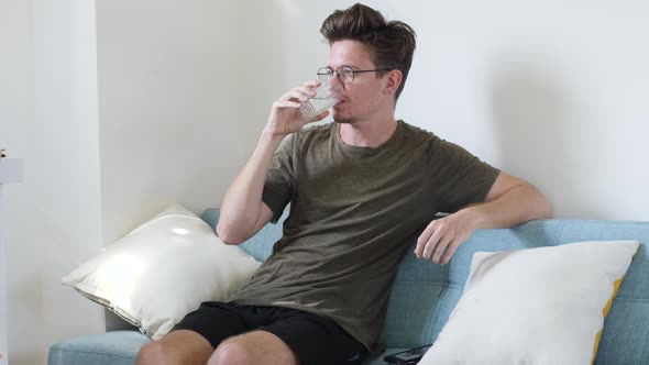 A Young European Man Drinks Clean Water From A Glass Sits At Home on A Bright Sofa in The Room