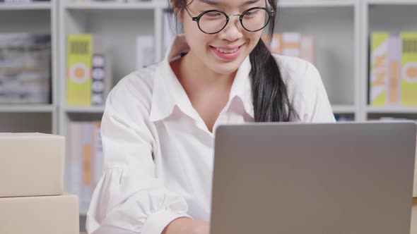 Front view of Happy young Asian woman seller wearing glasses keying information on laptop