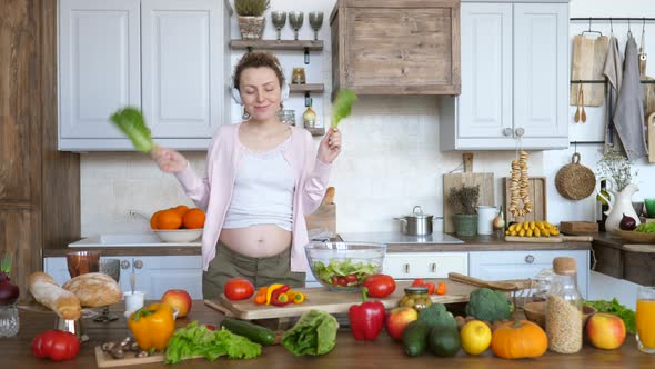 Happy Pregnant Woman Dancing With Green Salad Leaves In Kitchen While Cooking.