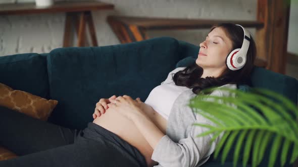 Pregnant Young Woman with Dark Hair Is Listening To Music Wearing Modern Wireless Headphones and