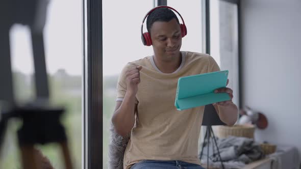 Middle Shot of Young Cheerful African American Man in Headphones Turning on Playlist on Tablet