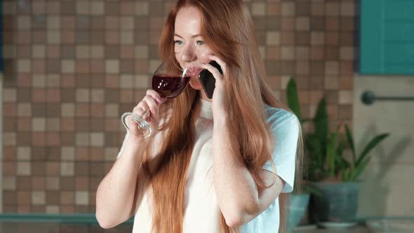 young red-haired woman with glass of red wine in kitchen talking on phone
