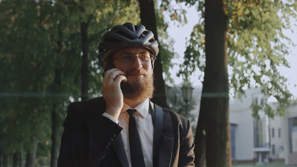 Business Man Wearing Bike Helmet Talks on the Phone Smiling in the Park