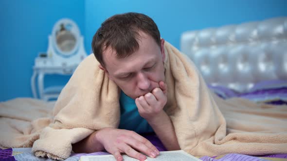 A man lies on the bed and reads a book, then covers his head with a blanket and falls asleep