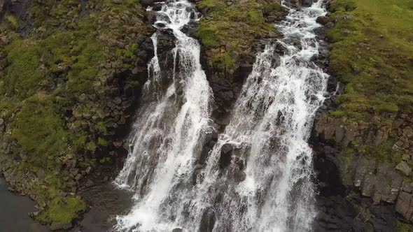 Aerial rising over Glymur waterfalls and river streaming down rocky cliff surrounded by verdant high