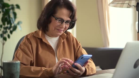 Ederly Woman Using Phone and Sitting at Table with Laptop in Apartment Room Indoors