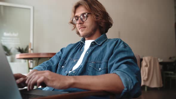 Smiling blond man in eyeglasses typing on laptop