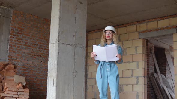 A Young Woman in a White Work Helmet and Denim Overalls and Goggles Stands at the Construction Site