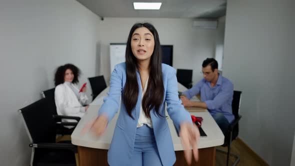 Emotional Asian Businesswoman Talking and Gesturing Looking at Camera with Colleagues Sitting at