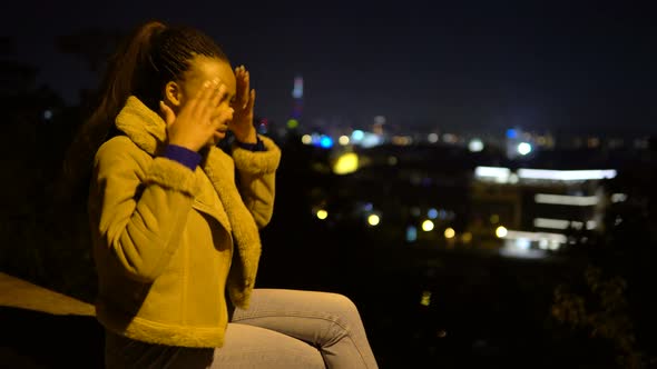 A Young Black Woman is Sick and Has a Headache As She Sits on a Low Wall in an Urban Area at Night