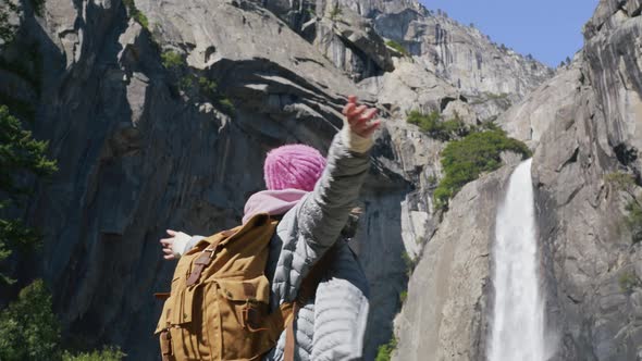 Young Woman with Tourist Backpack Looking at Scenic Waterfall on Top of Mountain