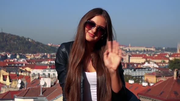 Young Beautiful Woman Sits on the Wall and Waves with Hand To Camera - City in the Background