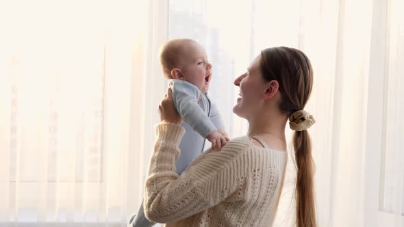 Happy Laughing Holding and Playing with Her Smiling Baby Son Against Sunset in Big Window